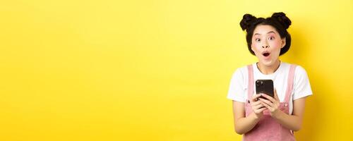 Excited girl with glam makeup and summer clothes, gasping amazed, checking out internet offer on mobile phone, standing on yellow background photo