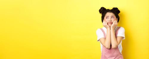 Sad moody asian girl, lean face on hands, looking left with sulking expression, standing in summer clothes on yellow background photo