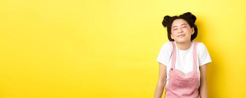 Romantic asian girl with cute pink makeup and summer clothes, close eyes and smiling dreamy, standing on yellow background photo