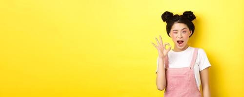 Alright. Confident asian female model with bright makeup and summer clothes, showing okay sign, winking and say yes, agree and approve, yellow background photo