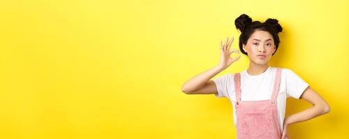 Sassy teenage asian girl with bright pink makeup, showing okay sign and nod in approval, approve product with serious face, not bad gesture, standing against yellow background photo