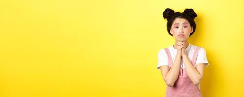 Hopeful asian stylish girl holding hands in begging sign, waiting with hope, asking for favour or help, standing on yellow background photo