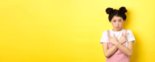Indecisive timid asian girl pointing sideways, showing left and right ways and sulking upset, standing sad on yellow background photo