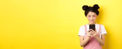 Stylish asian girl with glamour makeup using mobile phone, smiling at screen, standing on yellow background photo