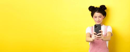 Stylish asian girl taking photo on smartphone, making with cellphone and smiling, standing on yellow background