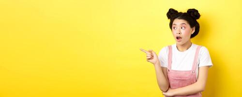 Intrigued asian beauty girl with makeup, looking curious at logo, pointing finger aside, standing in summer clothes against yellow background photo