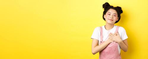 Beautiful teen asian girl in love, holding hands on heart and smiling touched, looking with affection, standing on yellow background photo
