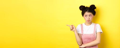 Upset frowning teenage girl sulking, pointing fingers left with jealous unfair face, standing disappointed on yellow background photo