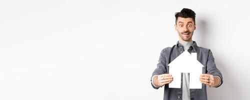 Real estate and insurance concept. Man look with excitement at paper house cutout, buying home, standing on white background photo