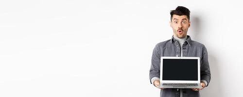 Excited man demostrated empty laptop screen, say wow and look at camera amazed, checking out online offer, showing webpage, standing on white background photo
