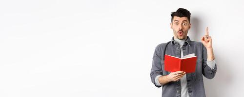 Excited man pitch an idea while reading planner or diary, holding red journal and gasping amazed, raising finger in eureka sign, standing on white background photo