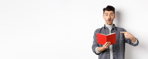 Excited guy read interesting thing in journal, pointing at planner and look impressed, standing against white background photo