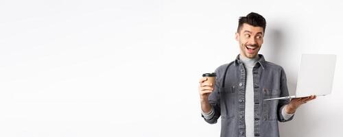 Cheerful young man working on laptop and drinking coffee, looking happy at computer screen, standing on white background photo