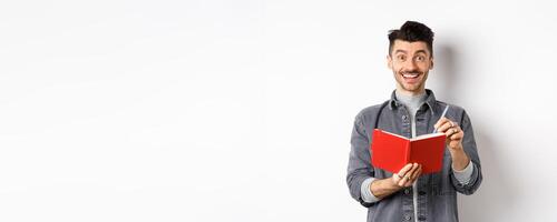 Creative young man smiling excited while taking notes, writing down ideas in planner, holding red journal and looking happy, standing against white background photo