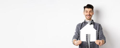 Real estate and insurance concept. Handsome young guy showing paper house cutout and smiling, searching for apartment, buying property, standing against white background photo