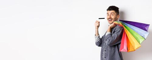 Cheerful guy going on shopping with credit card, holding bags on shoulder and smiling excited at camera, white background photo