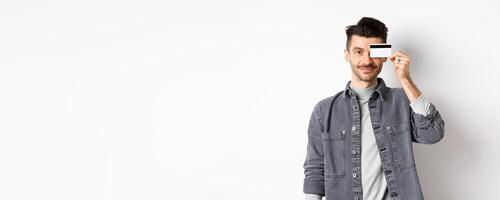 Handsome modern guy with moustache, standing in casual clothes and holding plastic credit card on eye, smiling pleased at camera, white background photo