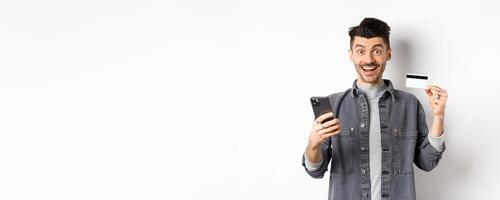 Online shopping concept. Excited man making purchase in internet, easy buying with plastic credit card and smartphone, smiling happy at camera, white background photo