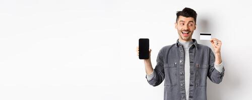 Excited handsome man showing plastic credit card and empty smartphone screen, advertising application, standing on white background photo