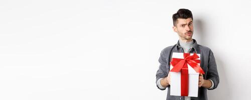 Surprised young man look with disbelief and hold surprise gift, raising eyebrow doubtful, being suspicious, standing on white background. Valentines day concept photo