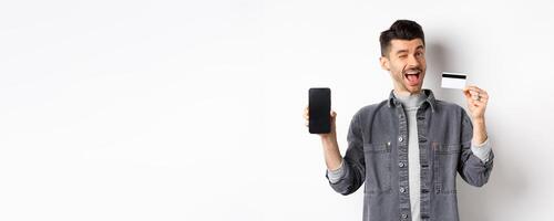 Cheerful handsome guy showing good deal, empty smartphone screen and plastic credit card, winking at you and smiling, recommending offer, white background photo