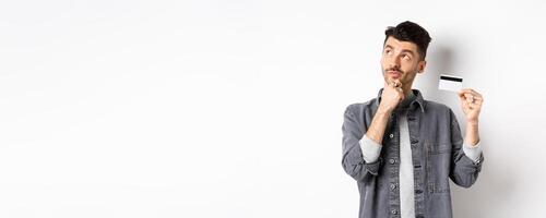 Thoughtful guy looking at upper left corner logo and holding plastic credit card, thinking about shopping, standing on white background photo