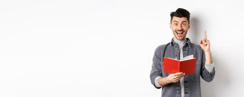 Excited guy pitching an idea while doing homework, raising finger and holding planner, standing against white background photo