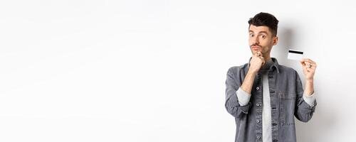 Thoughtful guy holding plastic credit card and thinking, looking intrigued at camera, touching chin while making choice, standing on white background photo