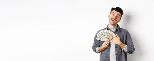 Happy smiling guy making money, hugging dollar bills with pleased relieved face, standing against white background photo