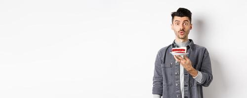 Handsome birthday guy blowing candle on cake and making wish, looking silly at camera, standing against white background photo