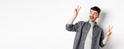 Funny young man showing v-signs and smiling at left side, looking at empty space with cheerful face expression, standing on white background photo