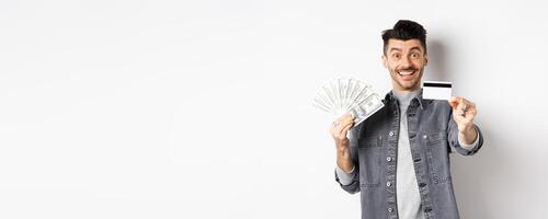 Image of handsome guy holding dollar bills but suggesting use plastic credit card, smiling friendly at camera, standing on white background photo