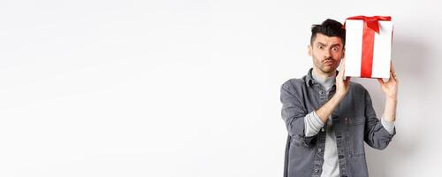 Intrigued young man looking confused at camera, trying to guess what inside gift box from lover, shaking present and stare puzzled, standing on white background. Valentines day concept photo