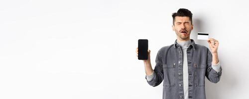 Sad crying man show empty mobile phone screen and plastic credit card, standing miserable on white background photo