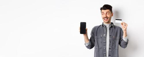 Handsome smiling man in denim jacket looking at empty smartphone screen and showing plastic credit card, good internet offer, standing against white background photo