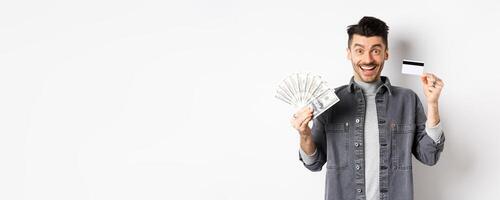 Excited guy holding plastic credit card and dollar bills, standing amused on white background photo