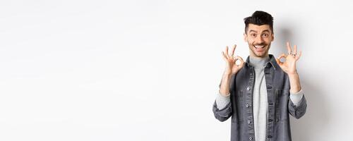 Happy guy say yes, smiling and showing okay signs, like good product, nod in approval and make satisfied face, praise excellent work, standing on white background photo