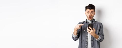Amazed young man showing cool thing on smartphone, looking and pointing at phone impressed, standing against white background photo
