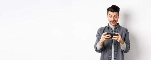 Excited guy playing game on mobile phone with focused face, tap on smartphone screen, holding cellphone horizontally, white background photo