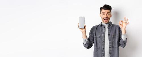 Excited smiling guy showing empty smartphone screen with okay gesture, recommending app or shopping offer, standing on white background photo