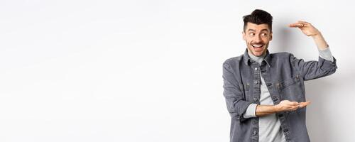 Excited guy looking at logo and showing big size thing, shaping large object and smiling amazed, standing in denim jacket on white background photo