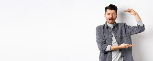 Sad man crying and showing big size object, shaping large thing and complaining, sobbing while standing on white background photo