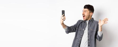 Shocked and confused man staring frustrated at smartphone screen, shouting from disappointment, standing against white background photo