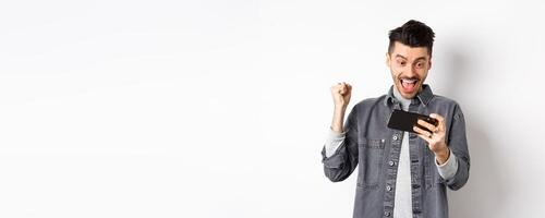 Happy man shouting yes, winning game on mobile phone and triumphing, achieve online goal, standing on white background photo