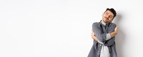 Happy romantic guy hugging himself with closed eyes and smile, loving own body, daydreaming about lover, standing on white background photo