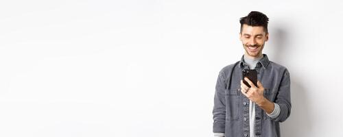 Handsome man reading message on phone and smiling. Guy holding smartphone and looking at screen, standing on white background photo