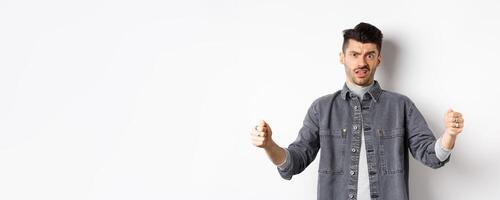 Skeptical young man showing big size object and frowning doubtful, standing reluctant on white background photo