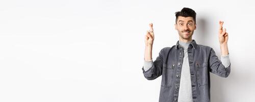 Hope dream come true. Positive and handsome guy making wish with fingers crossed, looking hopeful and smiling at camera, waiting results, standing on white background photo