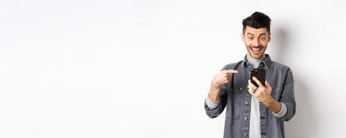 Excited handsome man showing cool news on phone, pointing at smartphone and smiling happy, standing against white background photo