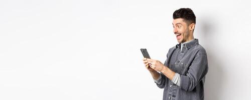 Profile portrait of excited handsome man reading message on phone, looking happy at smartphone screen, standing on white background photo